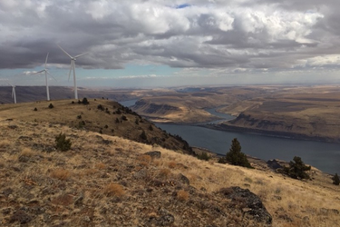 Landscape photograph of a high desert plateau. Three windmills spin in the distance beneath a stormy sky. The Columbia River is visible in the background.
